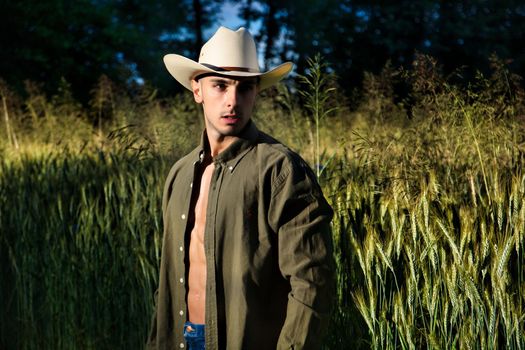 Portrait of sexy farmer or cowboy in hat with unbuttoned shirt on muscular torso, looking to a side, while standing next to hay field in countryside