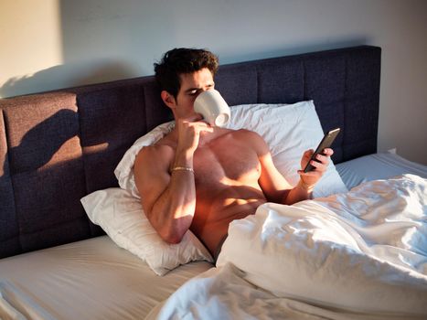 Naked young man with muscular body on bed with mug or cup in hand with coffee or tea talking on cell phone