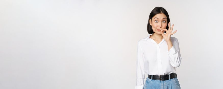 Mouth shut. Young asian woman seal lips, showing taboo dont speak, keep quiet sign, hiding secret, standing over white background.