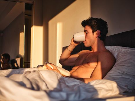 Young man lying in bed and reading a book with a cup of hot drink.