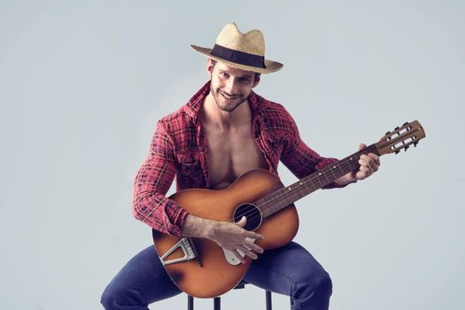 Studio portrait of handsome bearded man with unbuttoned shirt playing guitar over grey background, sitting on chair, looking at camera