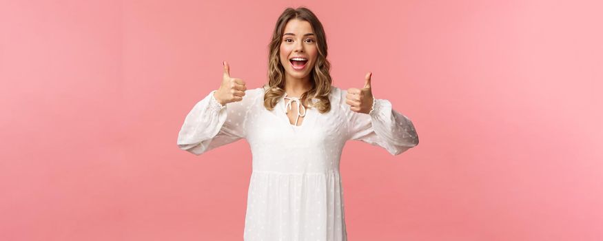 Portrait of feminine blond woman in white dress, show thumbs-up in approval, say yes, approve or agree, satisfied and recommend product, standing pink background in white dress.