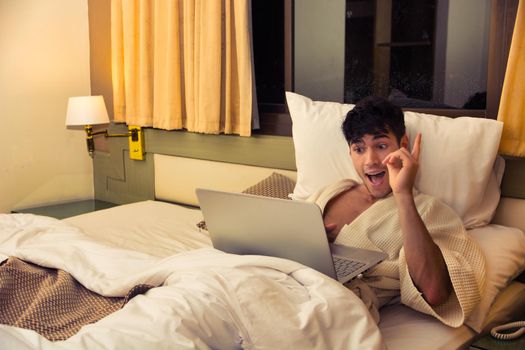 Attractive Young Shirtless Muscular Man with Serious Expression, Reclining Comfortably with Laptop on Bed Working on his Start-up Business - Young Male College or University Student Doing Homework, in Bedroom Looking at Camera