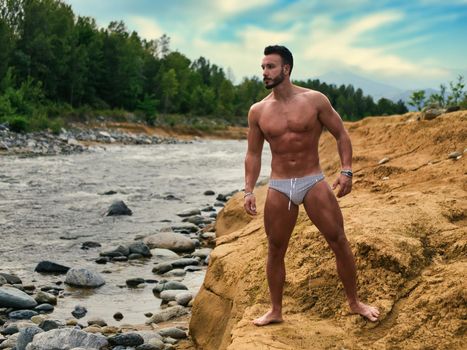 Attractive muscular shirtless young man in nature next to a river, wearing swimming suit, standing