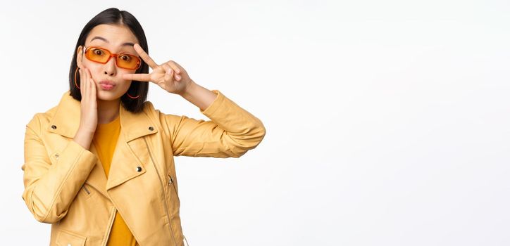 Portrait of stylish asian modern girl, wearing sunglasses and yellow jacket, showing peace, v-sign gesture, standing over white background, happy smiling face.