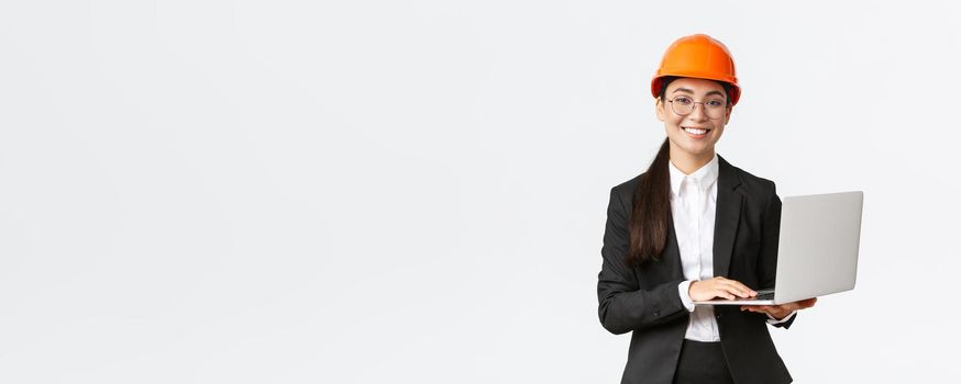 Smiling professional female saleswoman showing construction works on new house. Asian industrial engineer in safety helmet and business suit using laptop computer, enter data from factory.
