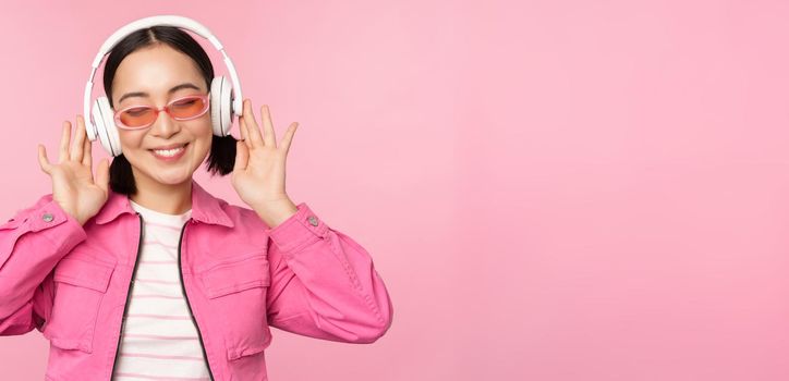 Dancing stylish asian girl listening music in headphones, posing against pink background.