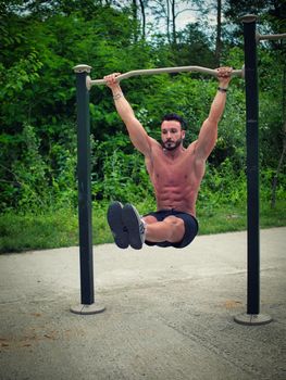 Attractive shirtless young man hanging from bar and working out in outdoor gym in city park