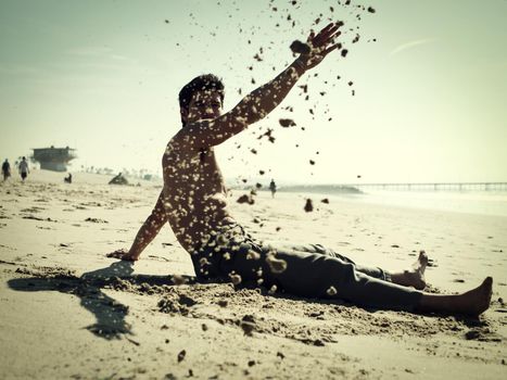 Handsome young man sitting on a beach playing with sand, shirtless wearing boxer shorts, showing muscular fit body