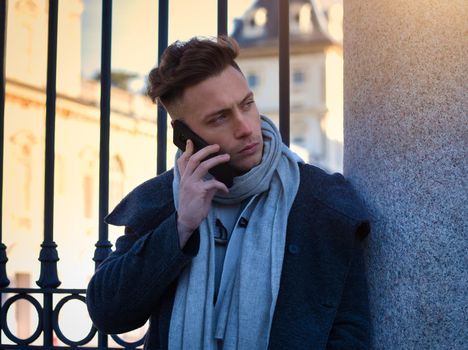 Handsome trendy man wearing white shirt standing and talking on cell phone, outdoor in city setting in day shot