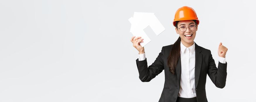 Successful winning asian female construction engineer, team lead architect achieve goal, holding house maket and fist pump in rejoice, celebrating victory in helmet and business suit.