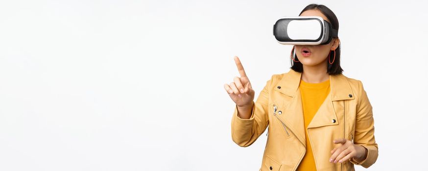 Young asian girl wearing VR glasses, looking amazed, using virtual reality headset, posing against white background.