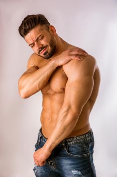 Muscular handsome man in jeans holding his shoulder with one hand and grimacing in pain in studio shot