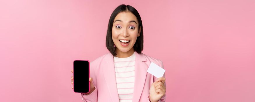 Enthusiastic asian businesswoman showing mobile phone screen and credit card, looking amazed at camera, standing over pink background.