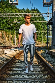 Attractive blond young man walking on railroad, wearing white shirt, looking away, at daytime