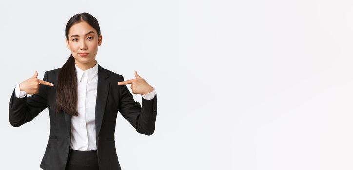 Professional asian businesswoman in suit pointing at herself as offering best deals. Proud and confident saleswoman show-off, suggest personal help or assistance, standing white background.