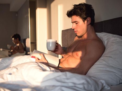 Young man lying in bed and reading a book with a cup of hot drink.