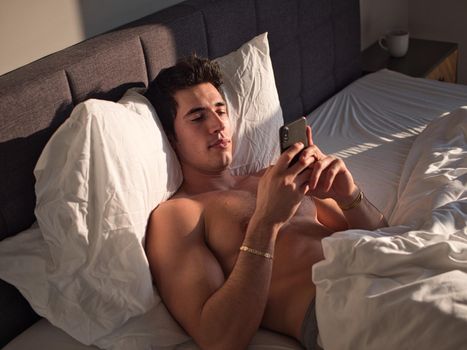 Handsome young man in bed typing on cell phone, sending text message or dialing number