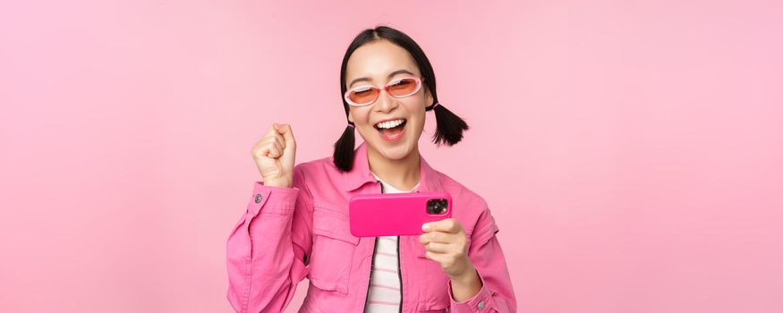 Happy smiling korean girl winning on mobile phone, looking at horizontal smartphone screen and rejoicing, achieve goal, celebrating, standing over pink background.