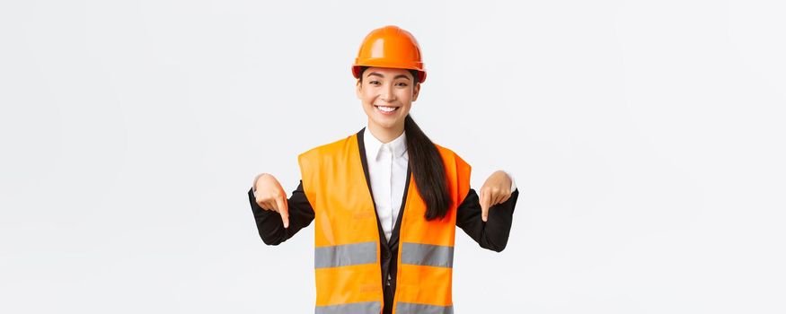 Smiling cheerful construction manager, asian female engineer in safety helmet and reflective clothing showing announcement. Woman project manager pointing down, standing white background.