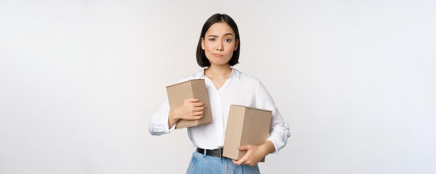 Complicated young asian woman holding two boxes, looking doubtful at camera, standing over white background puzzled.