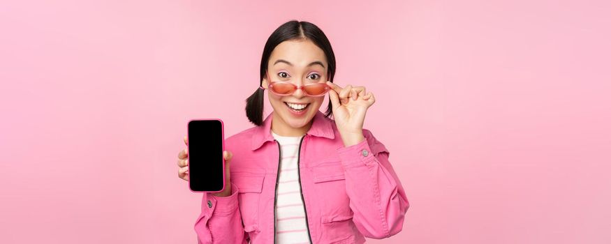 Portrait of asian girl showing mobile phone screen, reacting surprised, standing over pink background. Copy space