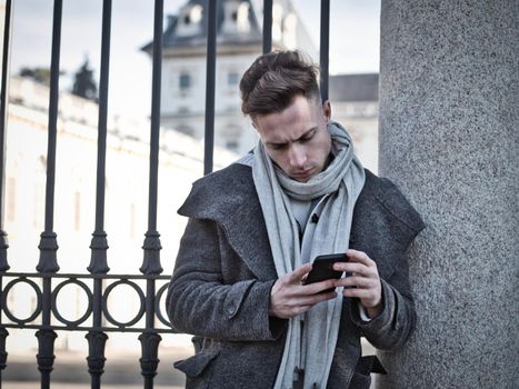 Handsome trendy man wearing cardingan standing and typing on cell phone, outdoor in city setting in day shot