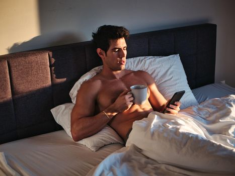 Naked young man with muscular body on bed with mug or cup in hand with coffee or tea talking on cell phone