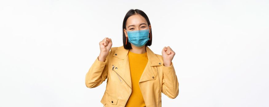 Covid-19 and people concept. Young asian woman in medical face mask, dancing happy in casual clothes, posing over white background. Copy space