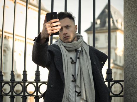 Portrait of young attractive man in winter coat using cell phone to take photo or selfie photo outside