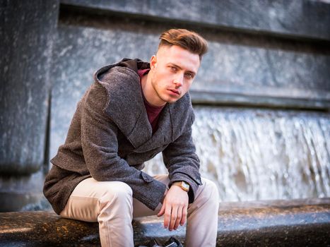Young man, a student, sitting by fountain, looking at camera in city setting in a winter day