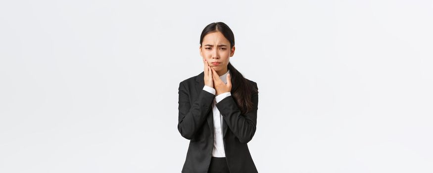 Female asian office manager in black suit having toothache at work. Troubled businesswoman holding hand on cheek as feeling pain in teeth, need appointment doctor, standing white background.