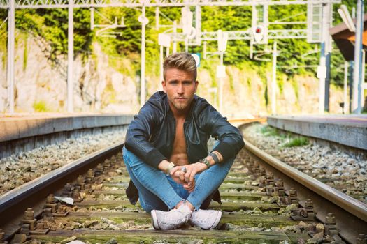 Attractive young man sitting on railroad, wearing black leather jacket, sneakers and jeans, looking at camera, at daytime
