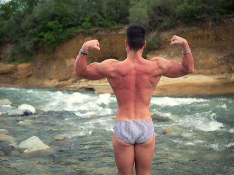 Attractive muscular shirtless young man in nature next to a river, wearing swimming suit, standing doing bodybuilding pose