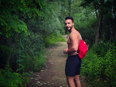 Handsome young man shirtless outdoors hiking on park path with backpack on shoulder