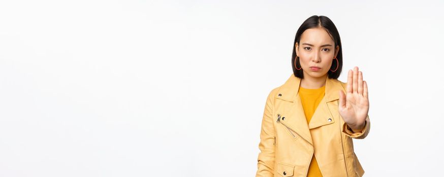 Serious and concerned asian girl extending palm hand, stop gesture, prohibit smth, forbid, standing over white studio background. Copy space