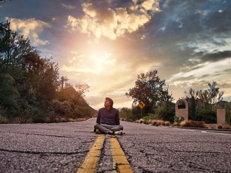 Attractive young man sitting in the middle of country road, with hoodie, looking away