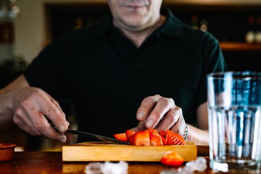 Detail of the hands of an experienced, concentrated and hard-working waiter, dressed in company uniform, black polo shirt, cutting strawberries for a cocktail on the counter of the nightclub. Preparing cocktails for customers. Warm atmosphere and dim light. Horizontal