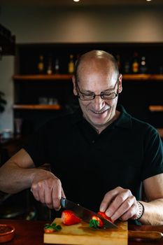 Professional bartender cutting fruit for a mojito. Waiter at the bar preparing drinks. Businessman in his restaurant with traditional ambience and backlighting, bottles of liquor in the background on shelves. Bald man with glasses preparing mojito.