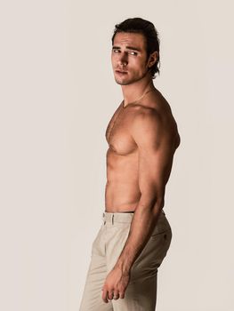 Three quarters, profile shot of handsome shirtless athletic young man in pants, looking at camera in studio shot