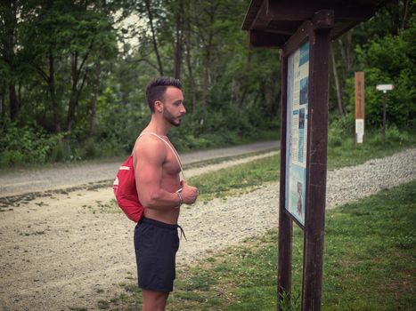 Handsome young man shirtless outdoors hiking on park path with backpack on shoulder