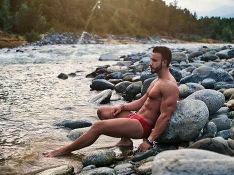 Attractive muscular shirtless young man in nature next to a river, wearing swimming suit, sitting on rock