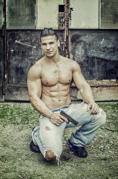 Menacing, muscular young man shirtless holding a handgun in his hand, outdoor