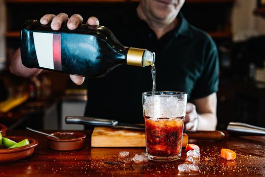 Detail of the hands of an experienced waiter, concentrated and precise, dressed in the company uniform, black polo shirt, pouring liquor into a crystal glass with ice on the counter of the nightclub. Preparing cocktails for customers. Warm atmosphere and dim light. Horizontal