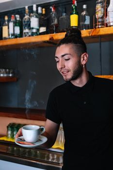 Young waiter with long hair tied back, concentrated and hard working, dressed in company uniform, black polo shirt, serving coffee in white cup Restaurant. Preparing coffee with milk. Warm atmosphere and dim lighting. Steaming cups of coffee. Vertical