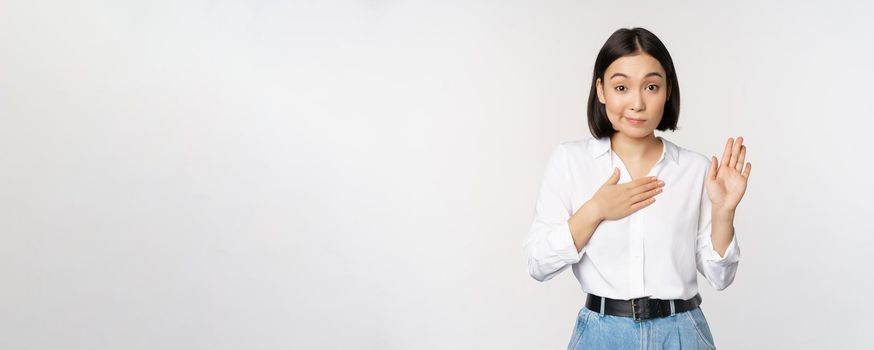 Image of cute young female office worker, asian girl student raising hand up and put palm on chest, name herself, introduce, making promise, standing over white background.