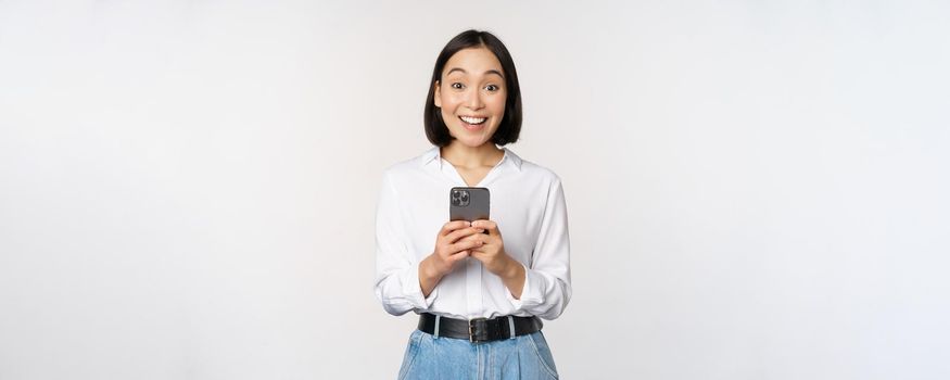 Excited asian woman smiling, reacting to info on mobile phone, holding smartphone and looking happy at camera, standing over white background.