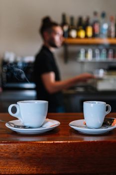 Detail shot of two white coffee cups on the bar counter. Waiting to be served to the customers. Background young waiter in black T-shirt. Warm atmosphere and dim light. Steaming cups of coffee. Vertical