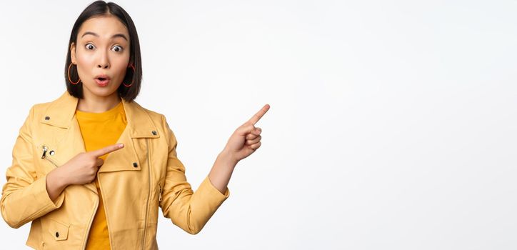 Enthusiastic asian girl pointing fingers right, showing advertisement and smiling, inviting to the store, standing over white background.