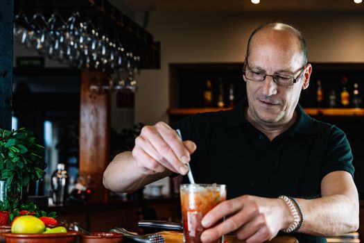 experienced waiter, concentrated and precise, dressed in company uniform, black polo shirt, stirring a cocktail in a crystal glass on the counter of the nightclub. Preparing cocktails for customers. Warm atmosphere and dim light. Horizontal